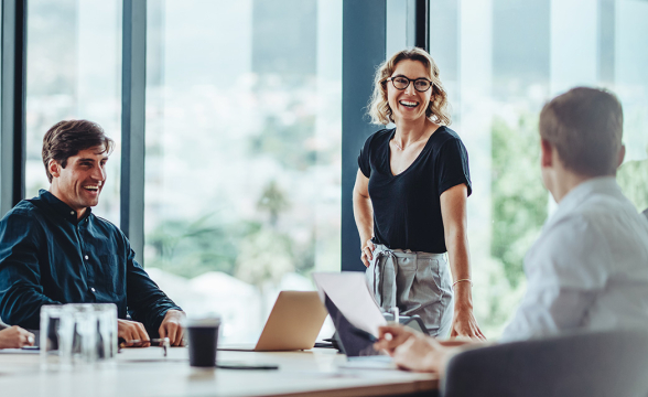 Eine Frau und zwei Männer sitzen in einem Besprechungsraum. Sie reden und lachen.