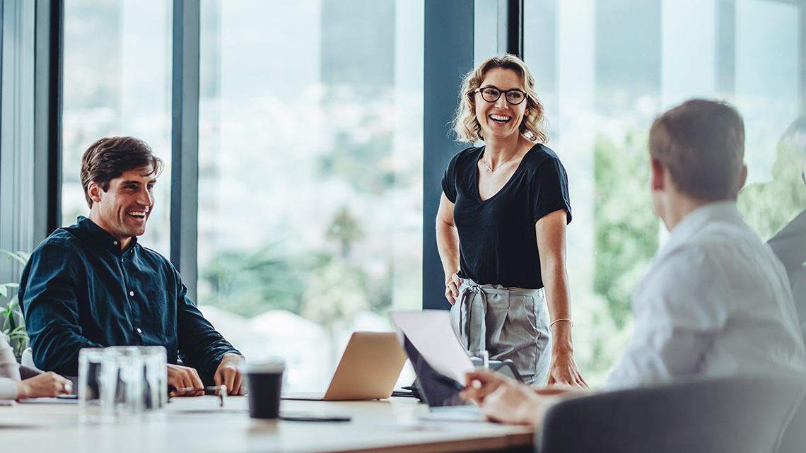 Eine Frau und zwei Männer sitzen in einem Besprechungsraum. Sie reden und lachen.
