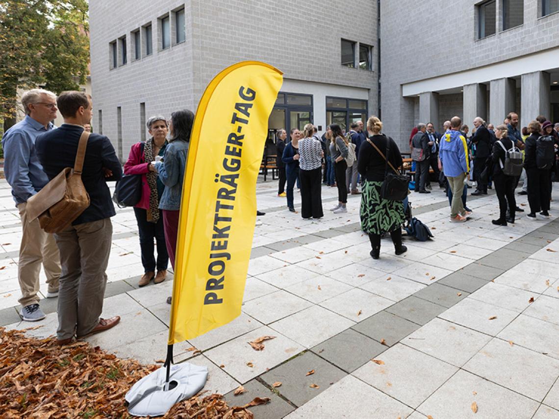 Viele Menschen gehen in eine Gebäude. Davor steht eine Flagge auf der steht: "Projektträger-Tag".