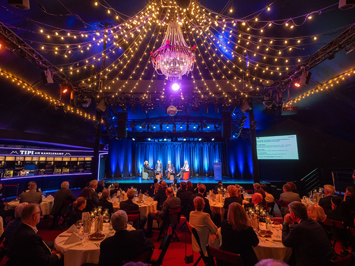 Ein Saal mit vielen Tischen, an denen Menschen sitzen und auf eine Bühne schauen. Auf der Bühne sprechen Menschen.