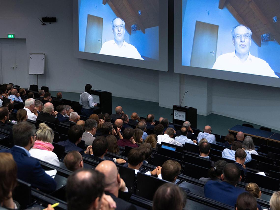 Ein Hörsaal gefüllt mit sitzenden Menschen. Sie sehen einen Mann auf einer Leinwand, der eine Rede hält.