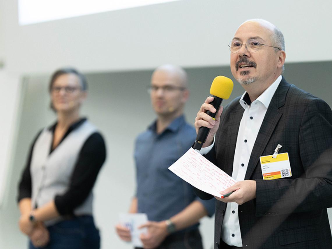 Ein Mann spricht in ein Mikrofon. Hinter ihm stehen eine Frau und ein Mann.