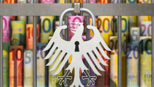 Symbolbild. Ein Vorhängeschloss in Form des Bundesadlers versperrt einen Käfig, in dem etliche Euroscheine stehen.