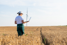 Farmerin mit weißer Cap, T-Shirt und Laptop auf Weizenfeld. Windkraftanlage im Hintergrund.